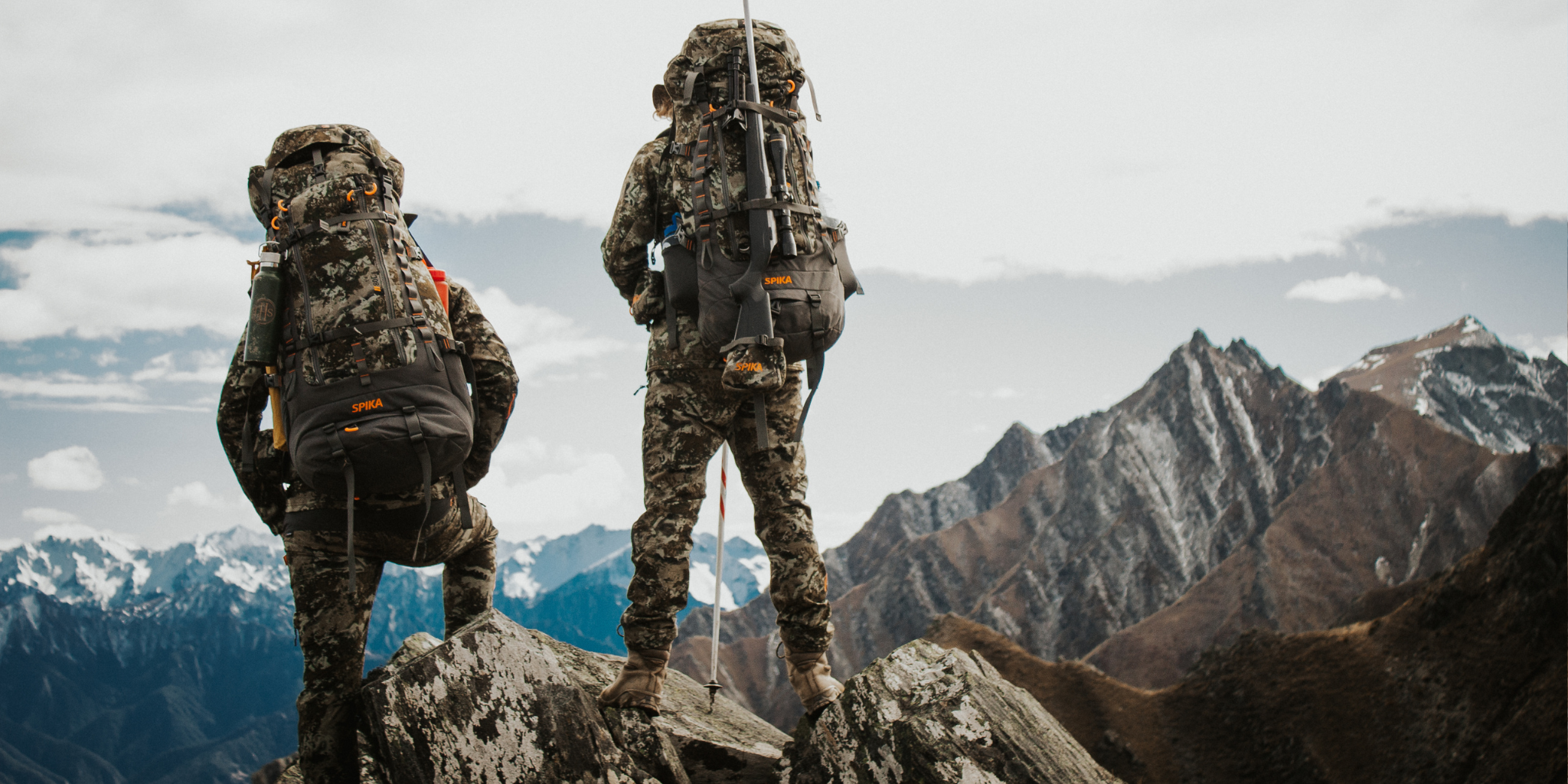 Two hikers in camouflage gear with backpacks standing on a mountain peak.
