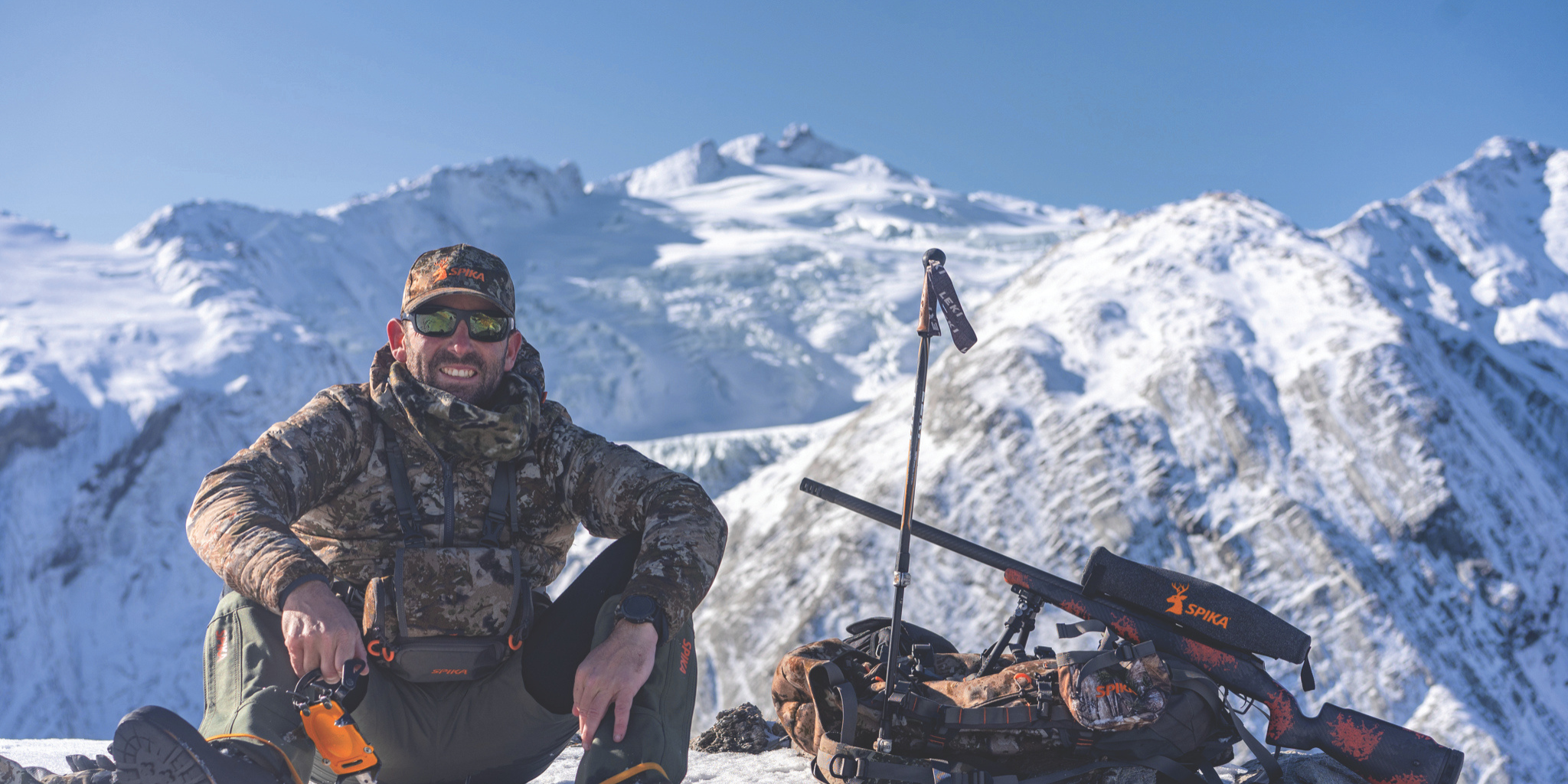 Spika Rep in camouflage gear with a rifle stand against a snowy mountain backdrop